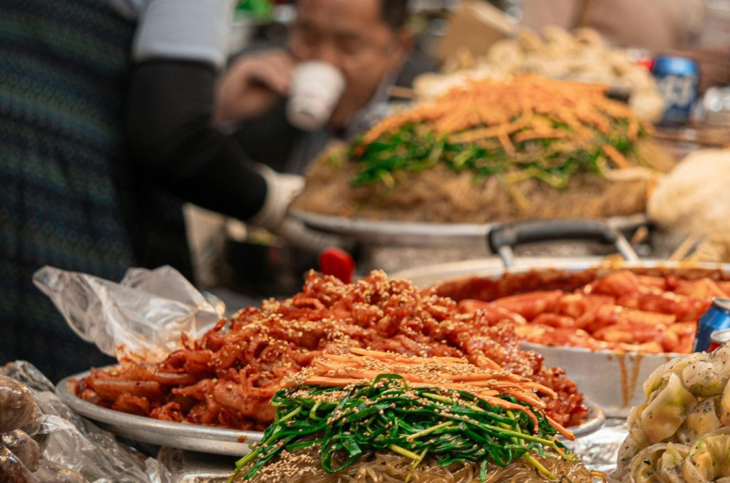 Colorful dishes at a bustling Asian market food stall with people in the background.